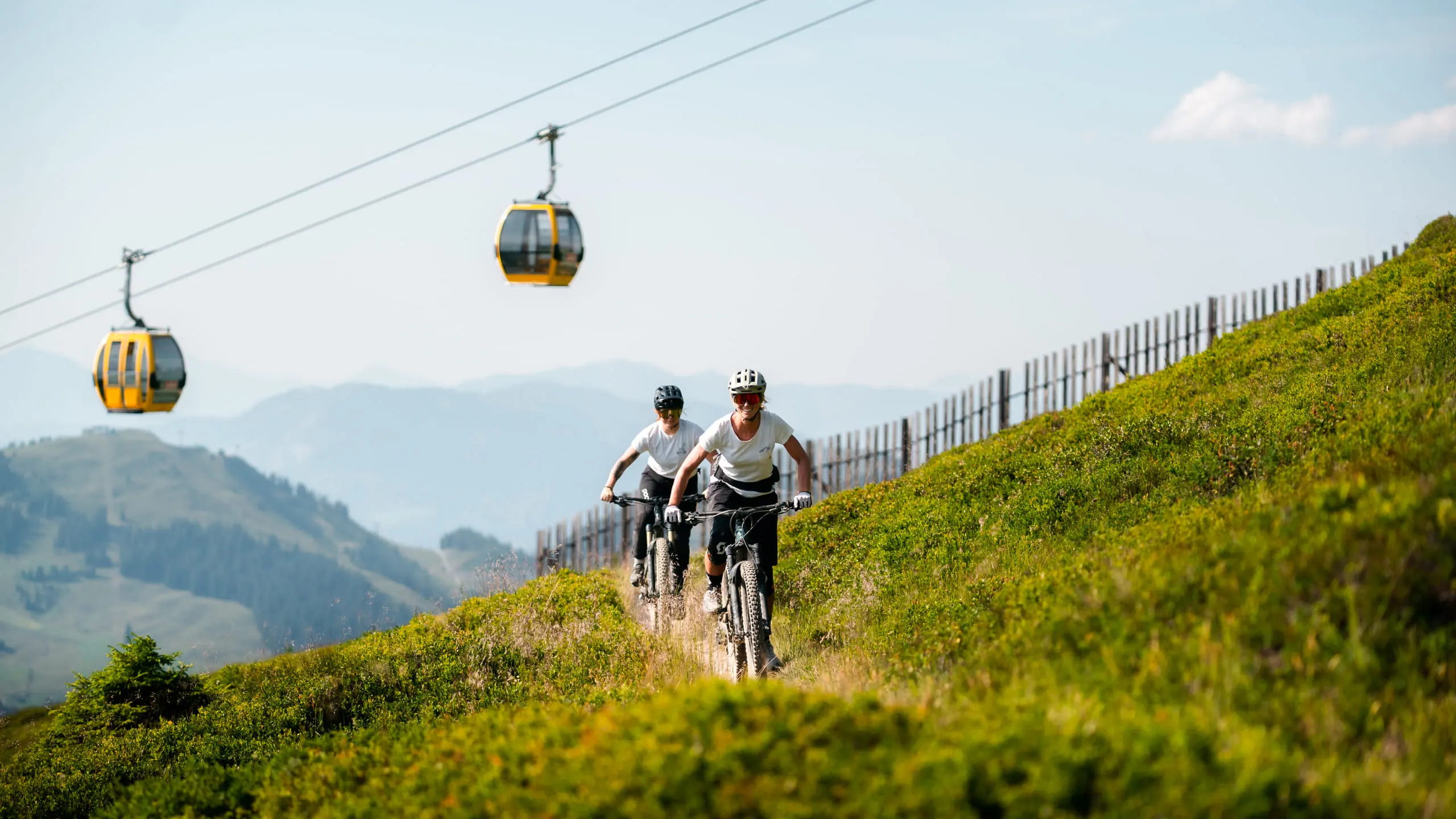 Zwei Radfahrer auf einem Hügel mit Seilbahngondeln im Hintergrund