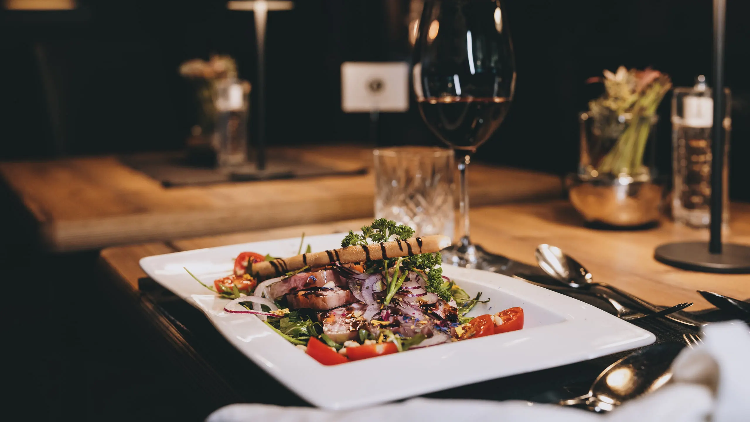 A gourmet dish on a white plate with vegetables and a wine glass in the background.