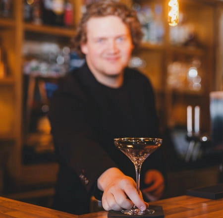 Bartender mit Cocktailglas vor einer Holzbar.