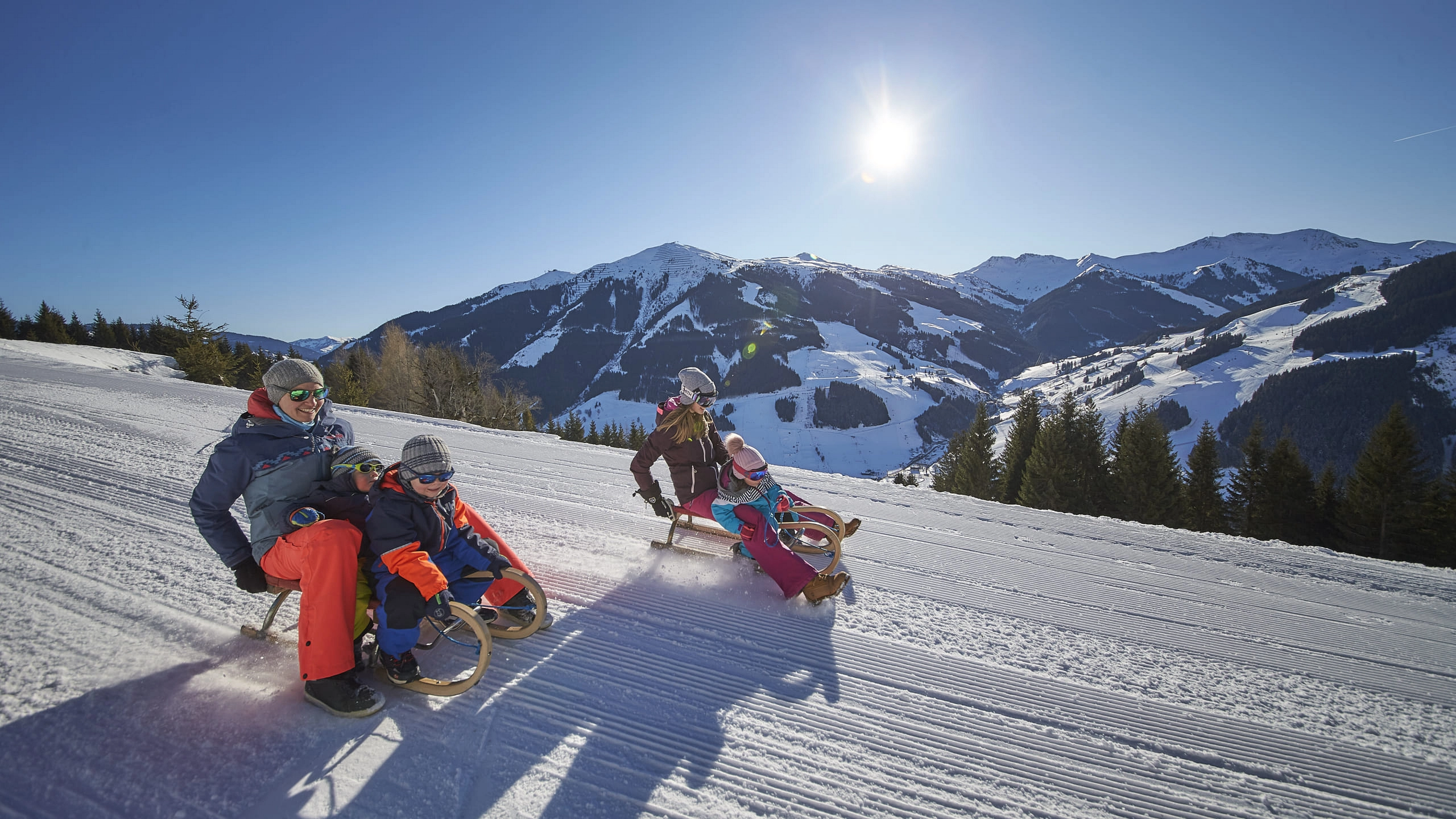 People tobogganing on a snowy slope with mountains in the background