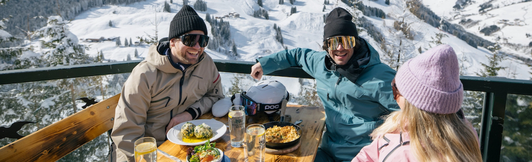 Drei Personen sitzen an einem Tisch mit Essen und Getränken in den Bergen im Winter.
