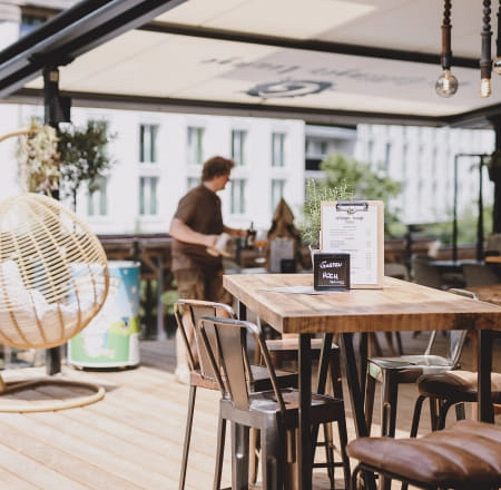 Outdoor-Bereich eines Cafés mit Holztischen und einem Stuhl aus geflochtenem Material.