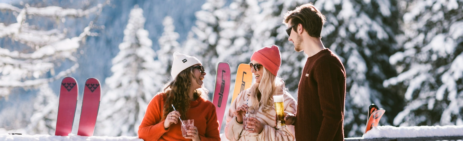 Drei Personen genießen Drinks im Schnee, Skiausrüstung im Hintergrund.