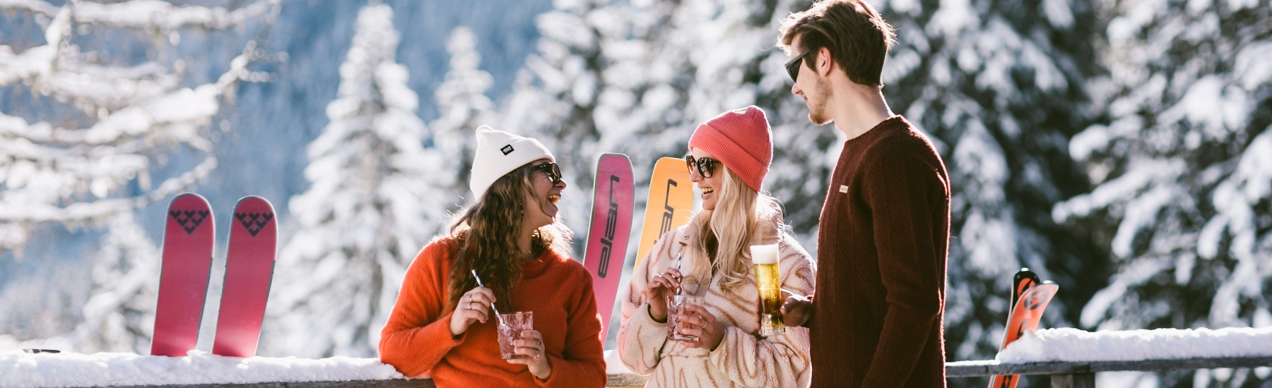Drei Personen mit Getränken stehen vor verschneiten Bäumen und Skiern.