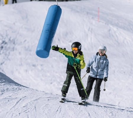 Ein Kind mit Skiausrüstung greift nach einem blauen Skilift, während eine Begleitperson zusieht.