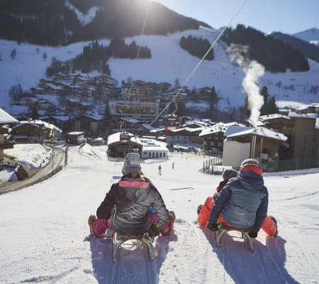 Drei Personen auf Schlitten blicken einen Hang hinunter in eine winterliche Landschaft.
